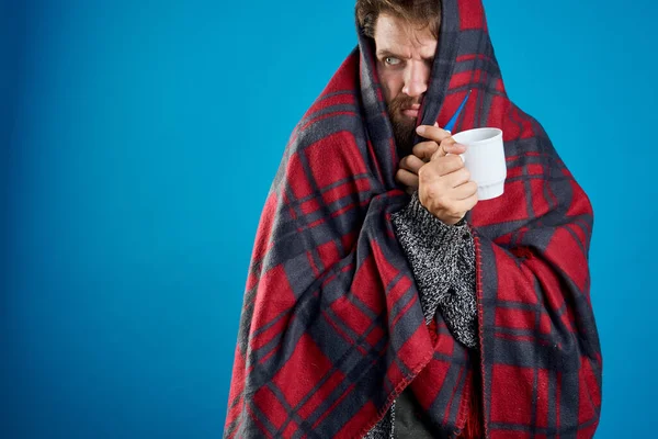 A man with a cup on a blue background and a warm blanket on his head — Stock Photo, Image