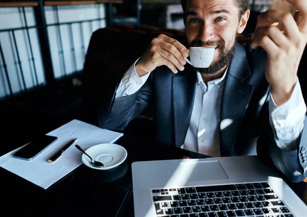 Uomo d'affari seduto in un caffè con una tazza di caffè tra le mani di fronte a una comunicazione tecnologia portatile — Foto Stock