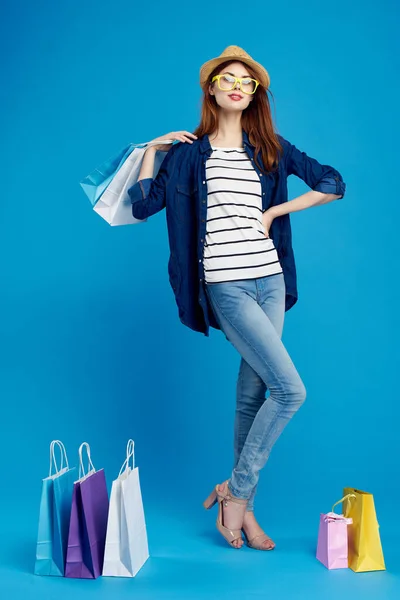 Mujer de moda es ir de compras con paquetes en un fondo azul en una camiseta a rayas y chaqueta, gafas en la cara —  Fotos de Stock