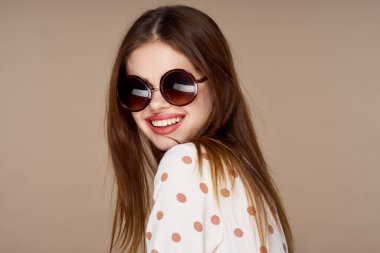  Güneş gözlüklü genç bir kadın bej arka planda yalnız poz veriyor.  