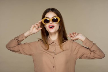  Güneş gözlüklü genç bir kadın bej arka planda yalnız poz veriyor.  