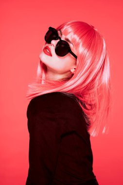 Pembe peruklu ve güneş gözlüklü kadın pembe arka planda poz veriyor.