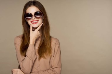  Güneş gözlüklü genç bir kadın bej arka planda yalnız poz veriyor.  