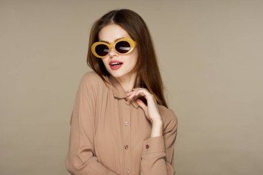  Güneş gözlüklü genç bir kadın bej arka planda yalnız poz veriyor.  