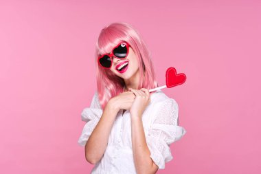 Pembe peruklu ve güneş gözlüklü güzel bir kadın pembe arka planda kırmızı kalp taşıyor.