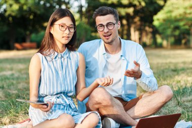 Dizüstü bilgisayarı ve tableti olan genç öğrenciler çimlerin üzerinde oturuyorlar. 