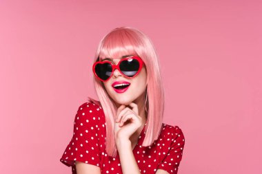 Pembe peruklu ve kalbi olan güzel bir kadın pembe arka planda güneş gözlüğü takıyor.