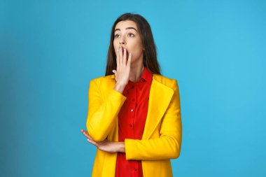 Stüdyoda sarı ceketli, kafası karışmış, mavi arkaplanı olan genç bir kadın..