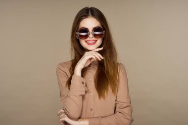  Güneş gözlüklü genç bir kadın bej arka planda yalnız poz veriyor.  