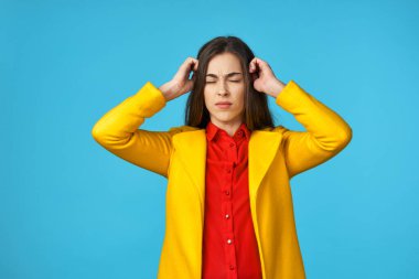 Sarı ceketli genç ve güzel bir kadın. Başı ağrıyor. Stüdyoda mavi arka planda..