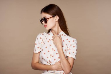  Güneş gözlüklü genç bir kadın bej arka planda yalnız poz veriyor.  