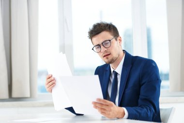 Gözlüklü genç yakışıklı işadamı masasında belgeler okuyor.