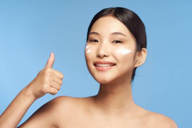 Genç Asyalı bir kadın mavi arka planda stüdyoda başparmağını gösteriyor. 