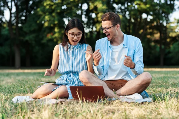 Dizüstü bilgisayarı ve tableti olan genç öğrenciler çimlerin üzerinde oturuyorlar. 
