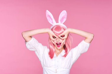 Pembe peruklu ve tavşan kulaklı genç bir kadın stüdyoda pembe arka planda gözlük yapıyor.