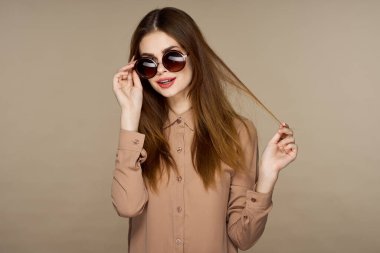  Güneş gözlüklü genç bir kadın bej arka planda yalnız poz veriyor.  