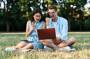 Dizüstü bilgisayarı ve tableti olan genç öğrenciler çimlerin üzerinde oturuyorlar. 
