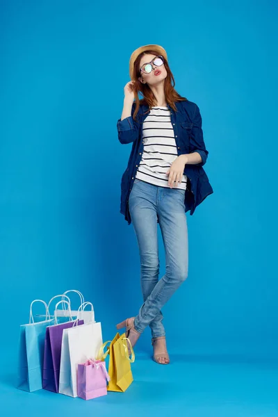 Mulher na moda está comprando com pacotes em um fundo azul em uma camiseta listrada e jaqueta, óculos em seu rosto — Fotografia de Stock
