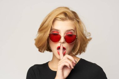 Sarışın, kısa saçlı, güneş gözlüklü siyah ceketli açık renk arka planlı.