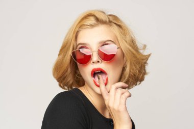 Sarışın, kısa saçlı, güneş gözlüklü siyah ceketli açık renk arka planlı.