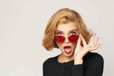 Sarışın, kısa saçlı, güneş gözlüklü siyah ceketli açık renk arka planlı.