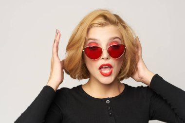 Sarışın, kısa saçlı, güneş gözlüklü siyah ceketli açık renk arka planlı.