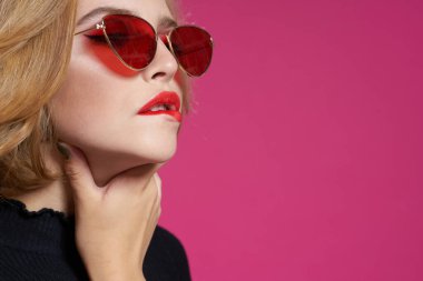 Koyu renk gözlüklü, parlak makyajlı, siyah ceketli, yakın plan pembe arka planlı güzel bir kadın.