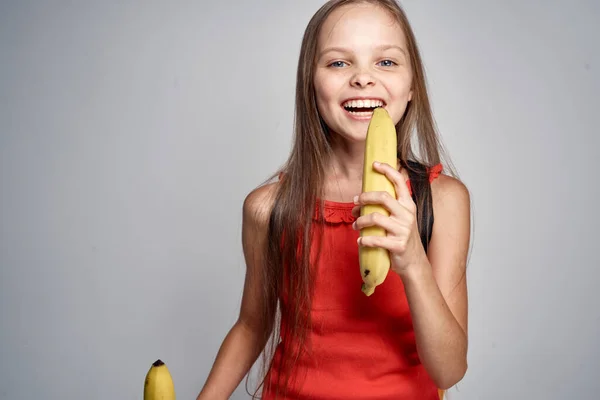 Little girl in glasses and a red dress with a banana in her hands light background — Stock Photo, Image