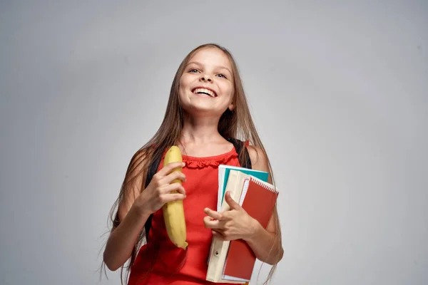 Bambina in occhiali e un vestito rosso con una banana tra le mani sfondo chiaro — Foto Stock
