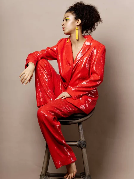 Una mujer en un traje brillante rojo con adornos de maquillaje brillantes se sienta en una silla descalza — Foto de Stock