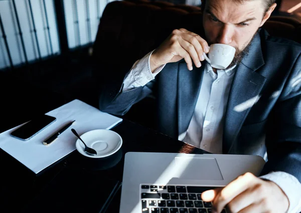 Homem de negócios sentado em um café com uma xícara de café em suas mãos na frente de uma comunicação de tecnologia de laptop — Fotografia de Stock