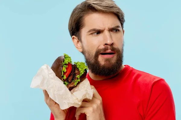 Hombre en una camiseta roja con una hamburguesa en las manos mira a un lado — Foto de Stock