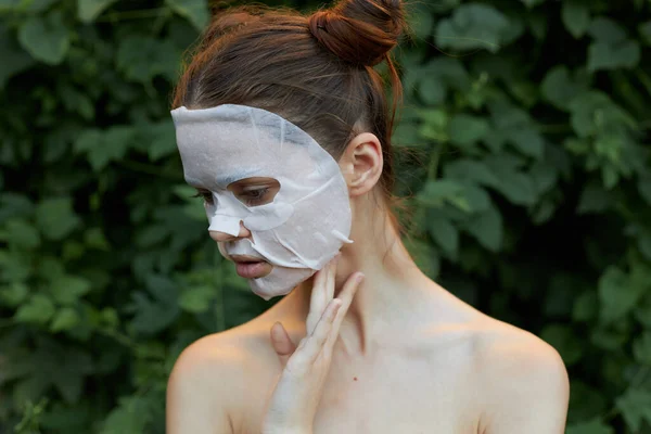 Retrato de uma menina máscara facial Olha para baixo e segura a mão perto de seu pescoço ombros nus arbustos no fundo — Fotografia de Stock