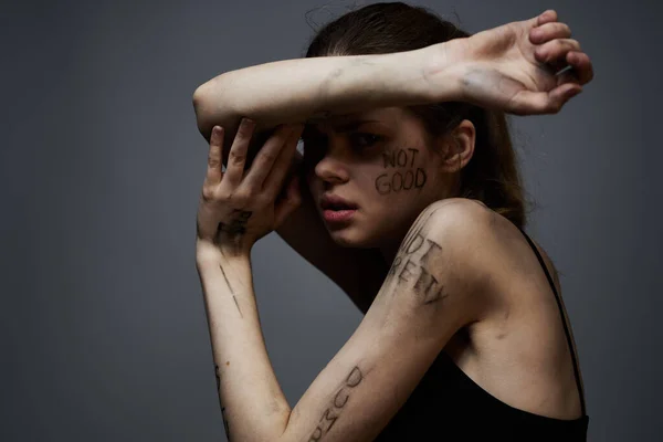 Mulher frustrada com inscrições ofensivas em seu corpo e em uma camiseta preta em um retrato de fundo cinza — Fotografia de Stock