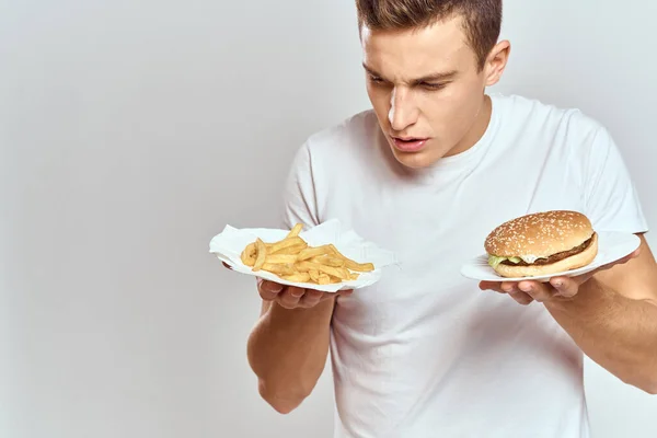 Ein Mann mit Pommes und einem Hamburger auf hellem Hintergrund in weißem T-Shirt in Großaufnahme — Stockfoto