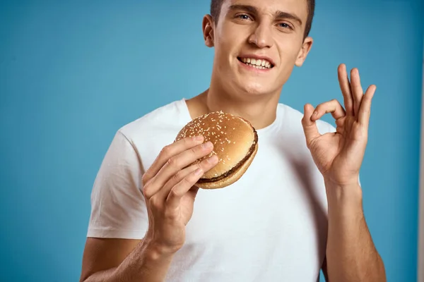 Hombre con hamburguesa y camiseta blanca fondo azul emociones gestos con las manos Copiar espacio — Foto de Stock