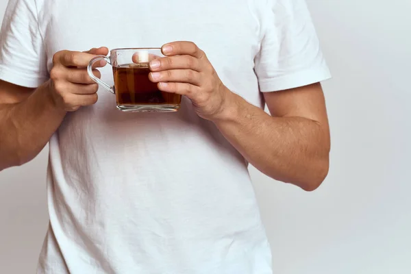 Um homem com uma bebida quente de chá em suas mãos em uma camiseta branca em um fundo leve vista cortada — Fotografia de Stock