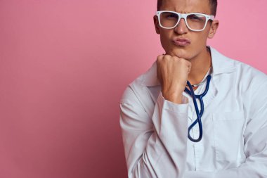 Pembe bir arka planda steteskop ve yüzünde gözlük olan tıbbi önlüklü bir erkek doktor.