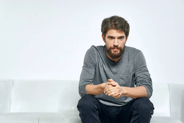 A lonely man in a sweater and trousers sits on a sofa on a light background