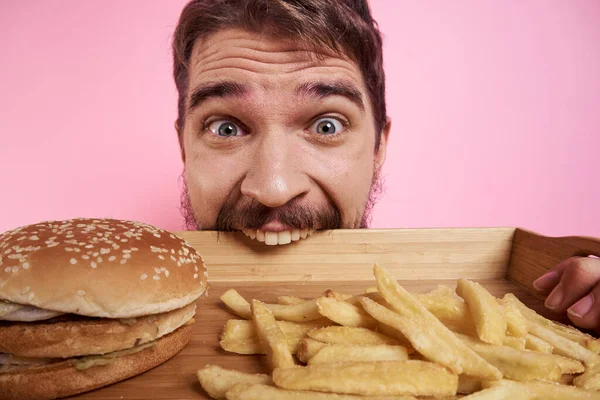 Cara agressivo com uma bandeja em suas mãos hambúrguer batatas fritas rosa fundo com fome olhar — Fotografia de Stock