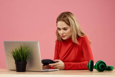 Dizüstü bilgisayarın önündeki masada oturan bir kadın elinde bir oyun platformuyla teknoloji kulaklığı çalıyor. 