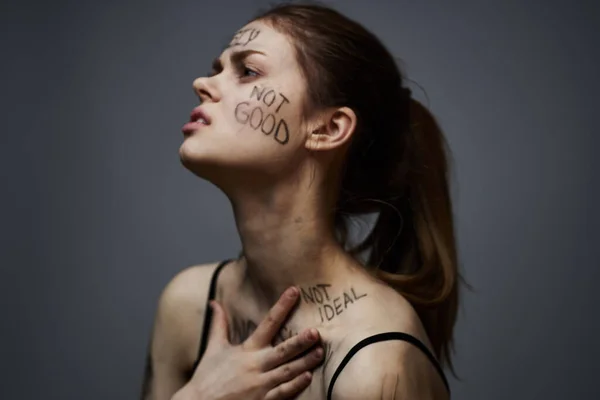 Ofendida mujer con inscripciones enojadas en su cuerpo se toca con las manos sobre un fondo gris agresión — Foto de Stock