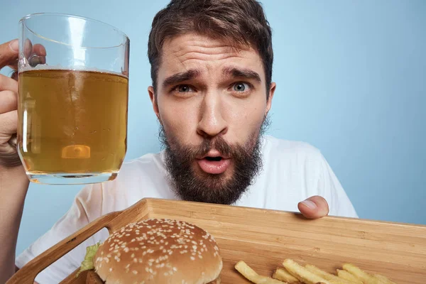 Homem alegre caneca de cerveja hambúrguer batatas fritas fast food dieta azul fundo close-up — Fotografia de Stock