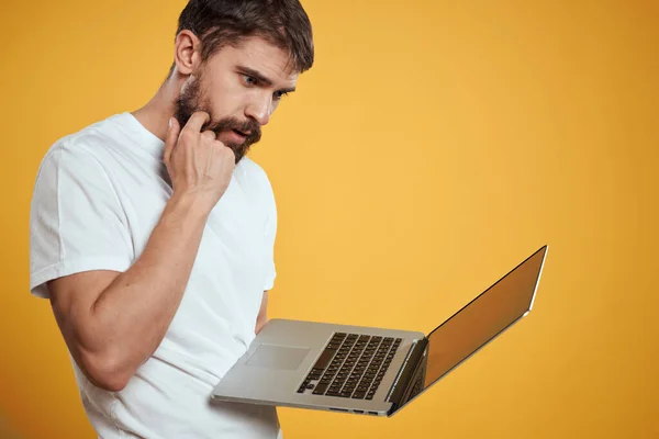 Un hombre con barba sostiene un portátil en su mano sobre un teclado de fondo amarillo monitor de nuevas tecnologías vista lateral — Foto de Stock