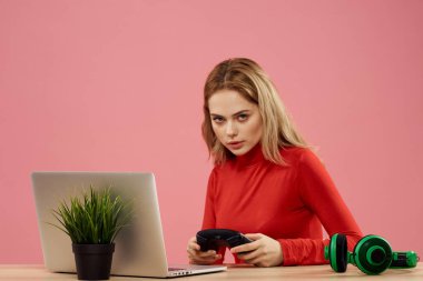 Dizüstü bilgisayarın önündeki masada oturan bir kadın elinde bir oyun platformuyla teknoloji kulaklığı çalıyor. 