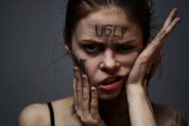 Gri arka planda kırıcı yazılar yazan üzgün kadın elleriyle kendine dokunuyor.