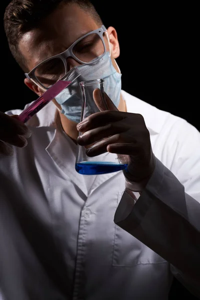 Assistant de laboratoire dans un masque médical et dans un manteau blanc avec un liquide dans une fiole et un produit chimique sur un fond noir — Photo