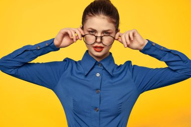 Mavi gömlekli, gözlüklü, yüzünde parlak makyajlı bir kadın elleriyle işaret ediyor.