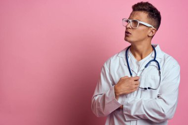Pembe bir arka planda steteskop ve yüzünde gözlük olan tıbbi önlüklü bir erkek doktor.