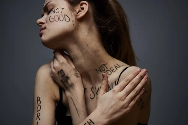 Ofendida mujer con inscripciones enojadas en su cuerpo se toca con las manos sobre un fondo gris agresión —  Fotos de Stock